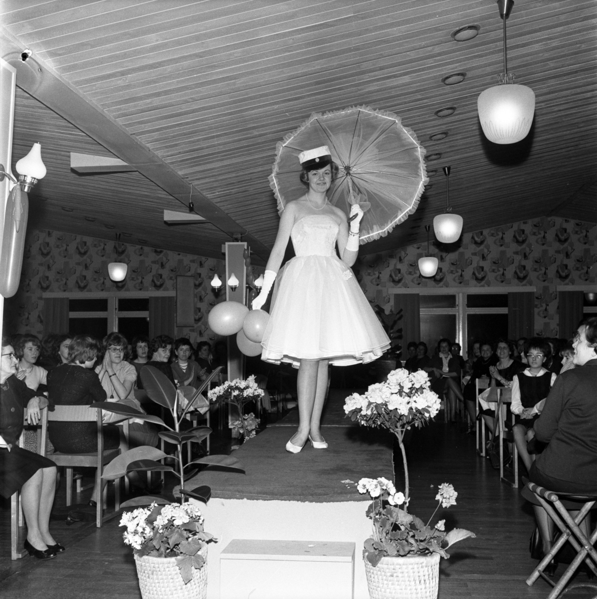 Modevisning i Huskvarna Folkets park våren 1961. På podiet går Gunnel Steén iklädd vit klänning, studentmössa och bär på ett parasoll. Medarrangör var Wera-magasinet i Huskvarna och konferensier var dess föreståndare Bitta Snygg.