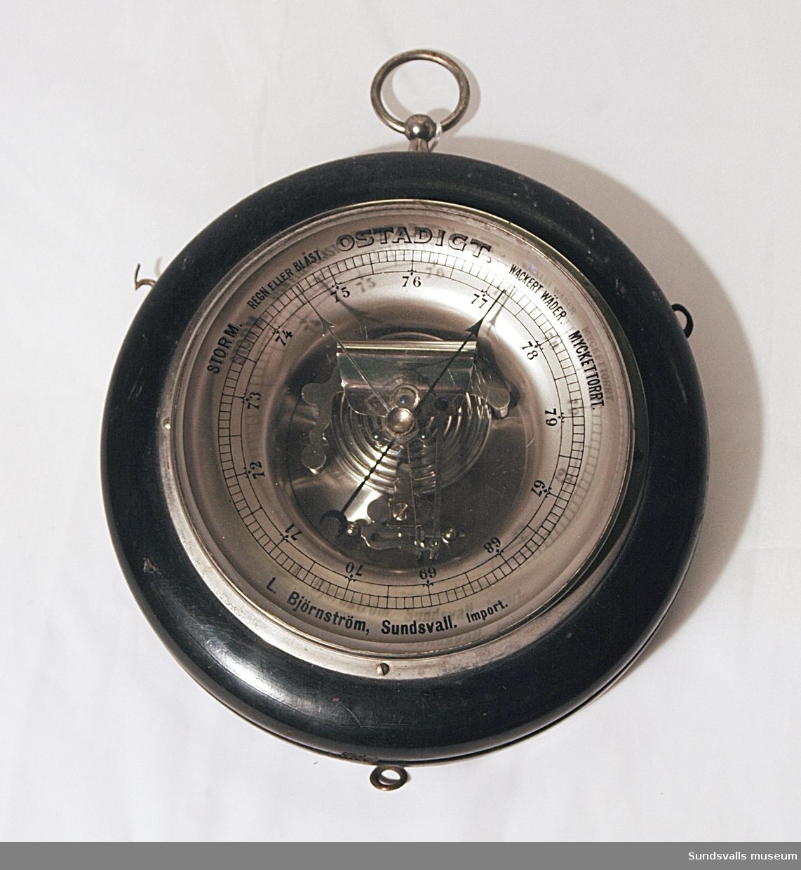 Barometer på svartmålad träplatta med upphängningsring i metall. På glaset finns text tryckt i svart.