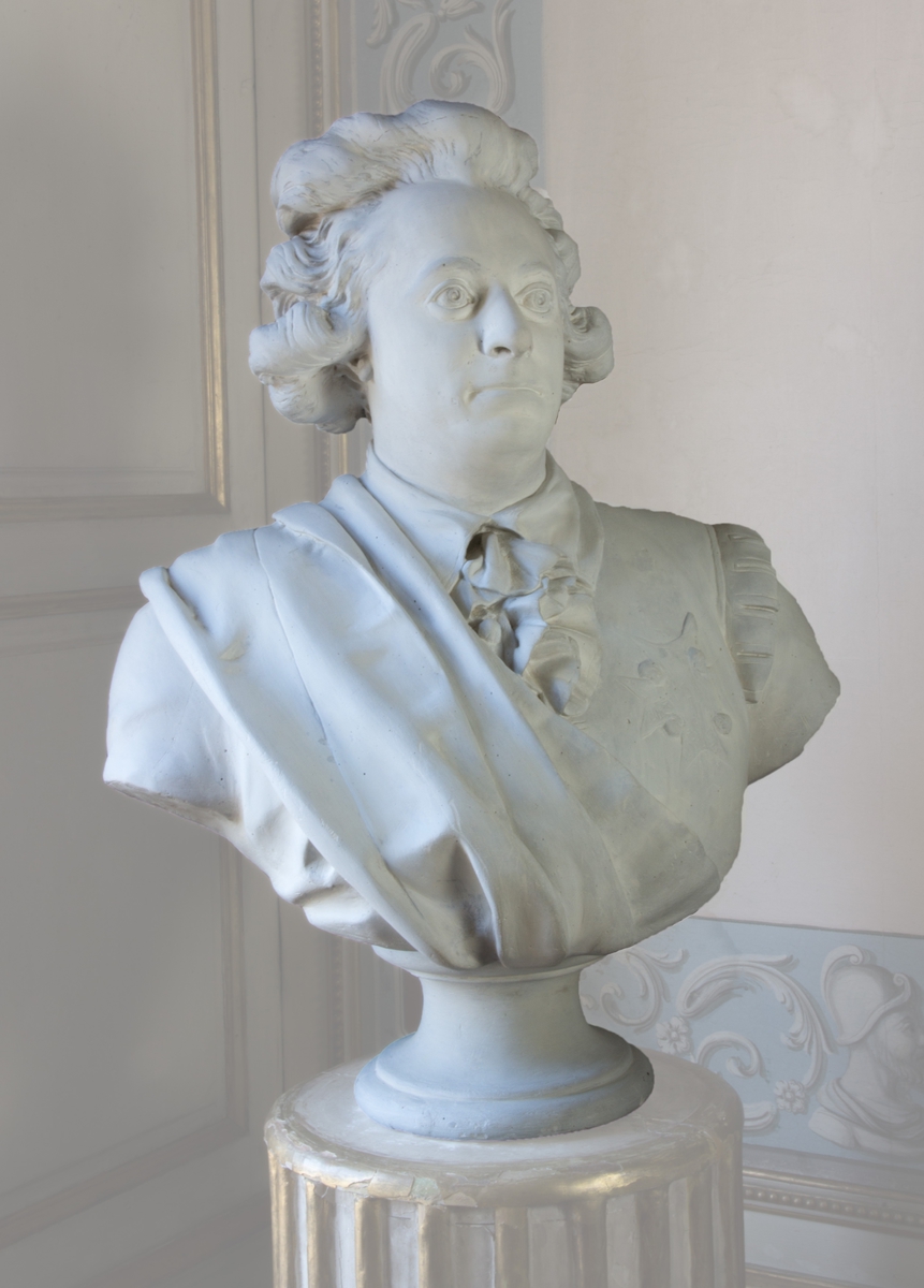 Byst av obehandlad gips, föreställande Gustav III i Svenska dräkten med band och kraschan, troligtvis tillhörande Serafimerorden, på pelarfot.