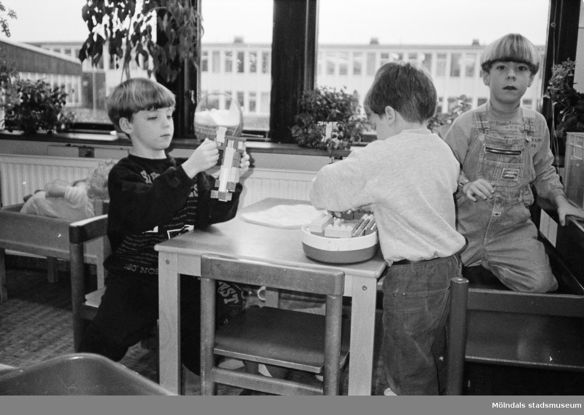 Tre pojkar sitter vid ett barnbord och leker med Duplo-lego. Vid sidan av dem står en docksäng. I bakgrunden syns fönster med Åbyskolans fasad. Katrinebergs daghem, 1992.