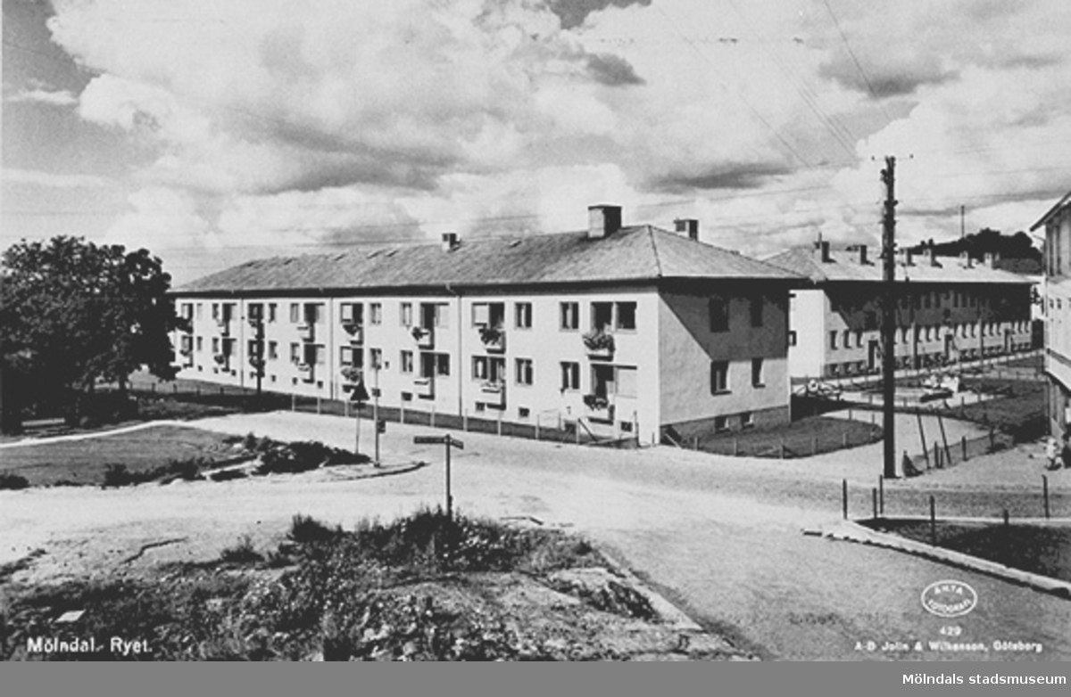 Man ser Rygatan 13-15. I bakgrunden ses Brunnsgatan 4 och längst till höger skymtar Rygatan 17. De två förstnämnda husen uppfördes 1949. Vykortet har producerats av A-B Jolin & Wilkenson, Göteborg. Kortet är märkt "Mölndal. Ryet. Äkta fotografi".