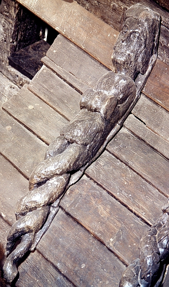 Triton med tjockt hår och stora mustascher. De förkrympta armarna är anslutna till en profilerad vulst som är placerad i midjehöjd och försedd med en bred skåra. Underkroppens övergång till dubbla fiskstjärtar markeras av bladliknande ornament.

Bakstycket är kraftigt och snidat i ett med skulpturen. Baksidan är försedd med hak eller urtag. Skulpturens profil är svagt S-formad.
Skulpturen är kraftigt sliten och fotpartiet delvis skadat.

Text in English: Triton with thick hair and large moustache. The arms are rudimentary. A torus sits at waist height and has a broad groove. The transition from the torso to the entwined fish tails is marked by a leafy ornamentation.

The backboard is robust and carved in one piece with the figure. The back side of the backboard was carved with hollowed out grooves which show that the sculptures were attached to a clinker-built surface. The profile is slightly S-shaped.
The sculpture has worn badly.