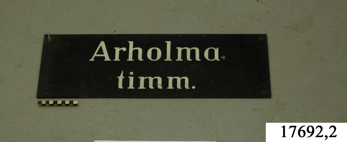 Rektangulär skylt, framsidan svartmålad, hål för upphängning. Vit schablonmålad text: " Arholma timm."