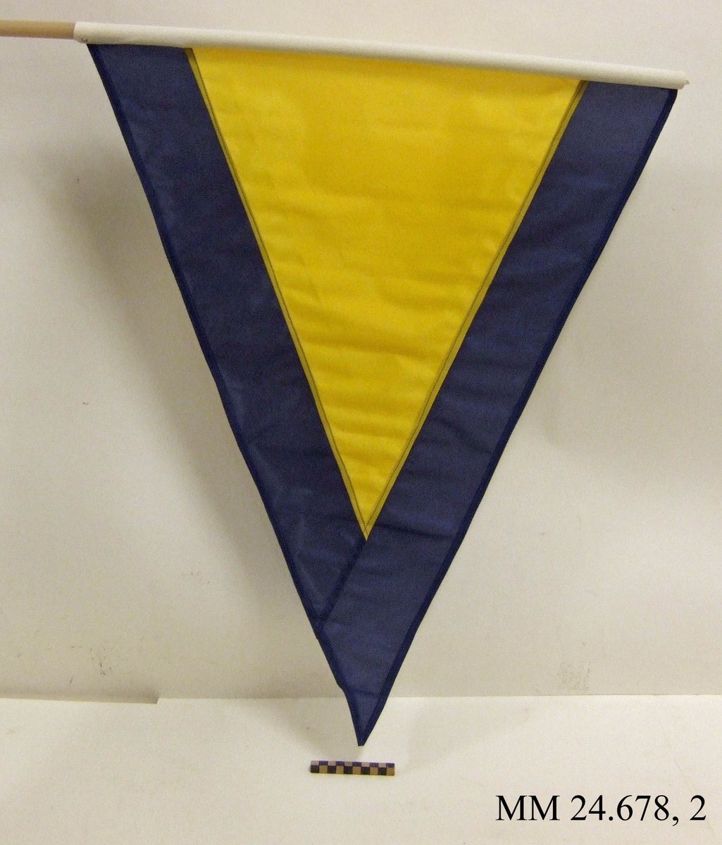 Fartstandert av nylon på rund stång av plast. Även standerten Alfa. Trekantig form, gul med mörkblå kant ca 120mm. Vitt lik trätt på plaststav och skruvat fast. Flaggan ligger i påse tillsammans med tre andra flaggor, sk. fartflaggställ M 8320-034010.