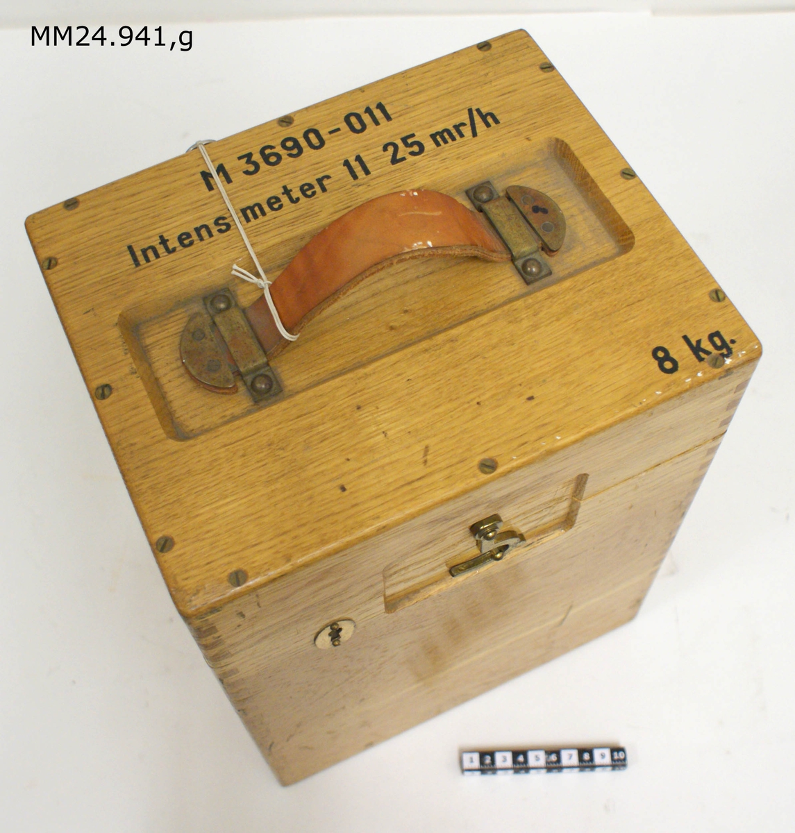Rektangulär låda i ljust trä med lock. På framsidan hasp och lås. På locket handtag i läder samt följande text:

M3690-011
Intensimeter 11 25 mr/h
8 kg

Inuti lådan två fack samt spännrem att fästa platspåsanr (e) i.