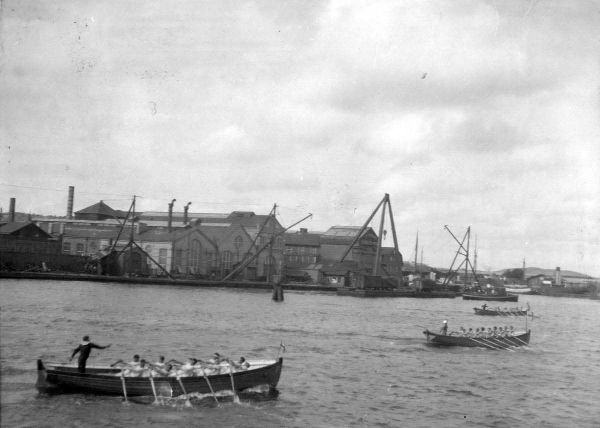 Män tävlar i rodd fördelat på tre båtar. I bakgrunden syns Karlskronas hamn med bebbyggelse.