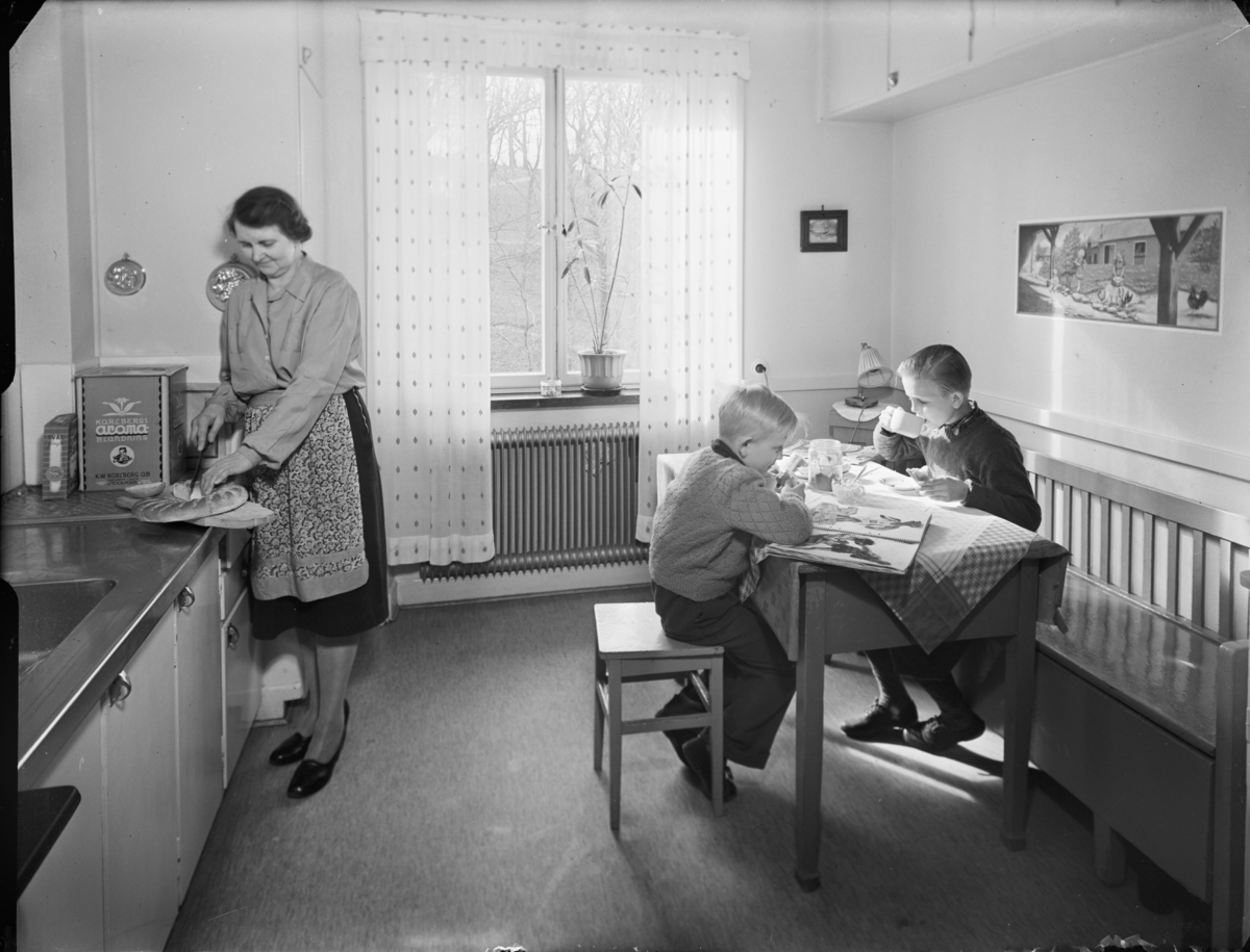 "Husmor med barn i hemmiljö"
Kök med en kvinna och två barn som äter mellanmål
Interiör