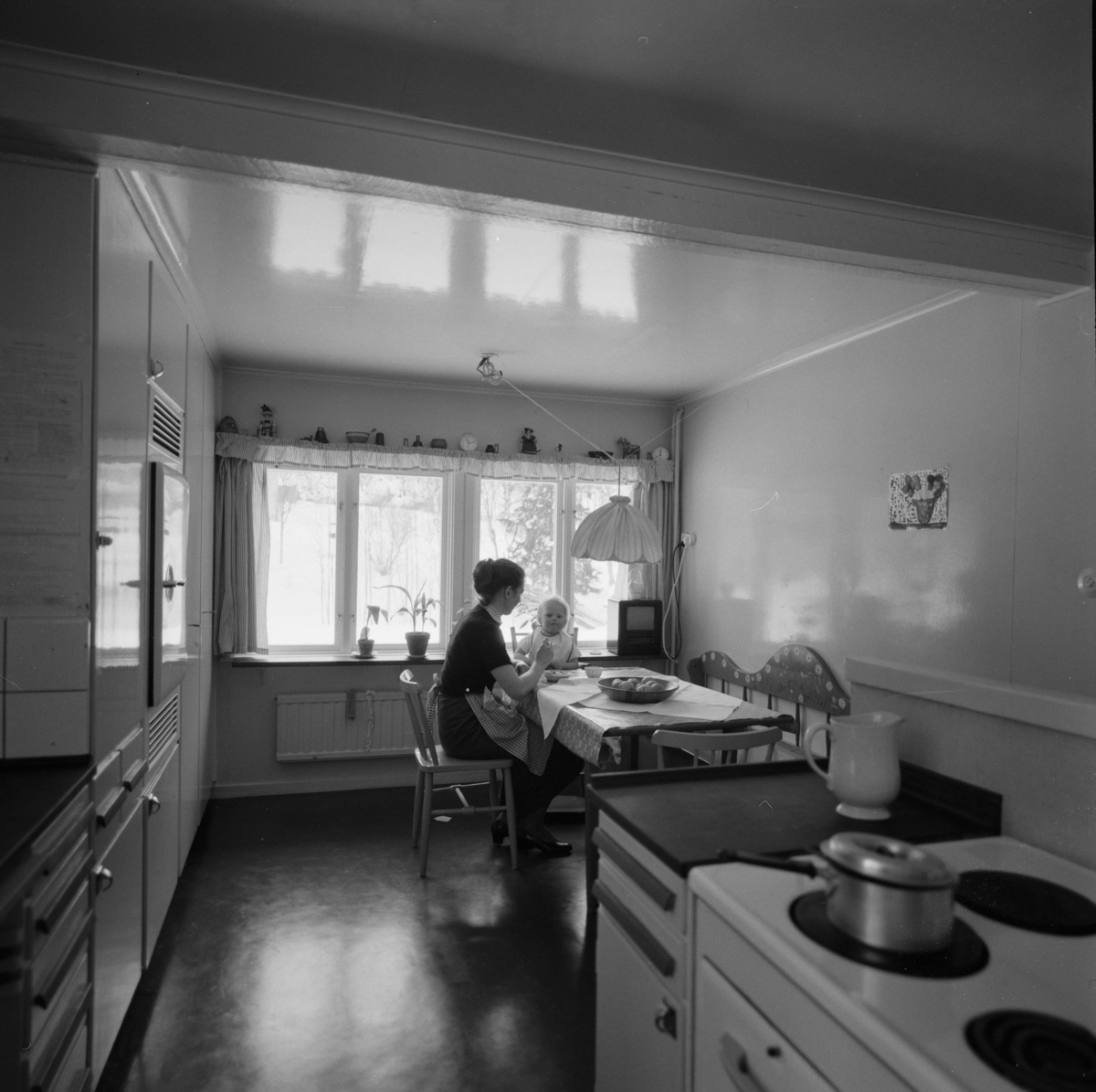 villa Ahlgren
Interiör av kök. Kvinna med barn i motljus mot fönster i fonden