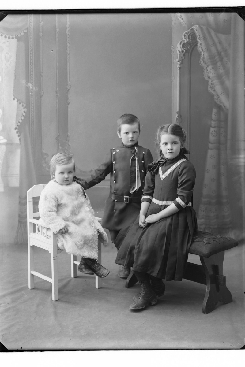 Studioportrett i helfigur av tre barn i finklær. Gutten i midten har på seg 17. maisløyfe.