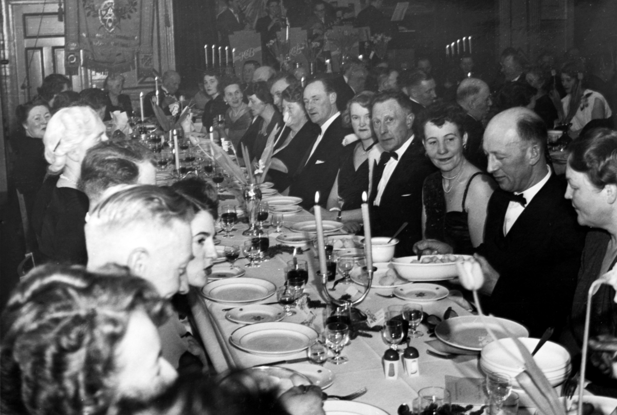 Festkledte kvinner og menn ved dekkede langbord. Bildet kan være tatt i forbindelse med 50-årsjubileet til Harstad Håndverkerforening, i 1952.