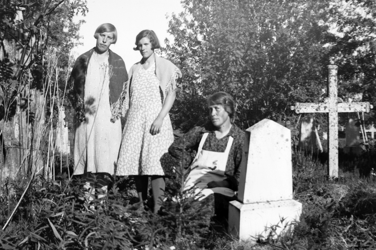 Tre kvinner ved en gravstein. Vegetasjon og trekors i bakgrunnen.