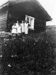 To kvinner og tre jenter står foran en liten tømmerstue med 