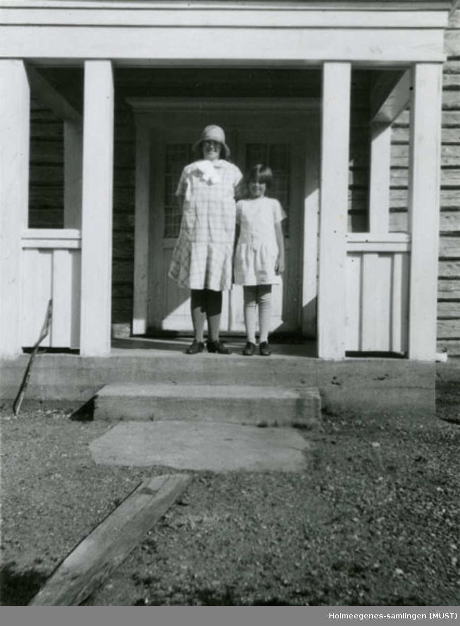 To jenter står på en overbygd trapp på et tømret hus. Antatt samme hus som ST.K.HE 2007-011-0120. Se også ST.K.HE 2007-011-0111 til -0126.
