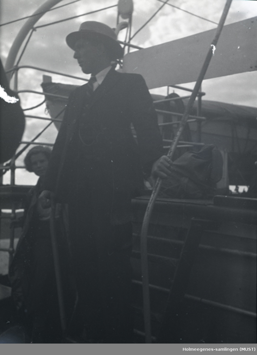 Mann med dress, hatt og stokk ombord på en båt. En kvinne titter frem bak ham.