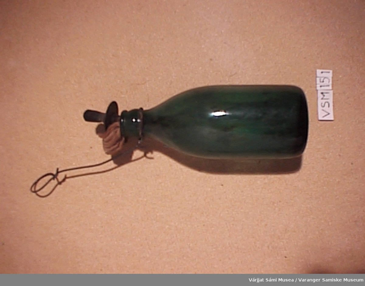 Grønn glassflaske med veik av tøy. Veikholderen av metall og flaskeopphenget er av metall.