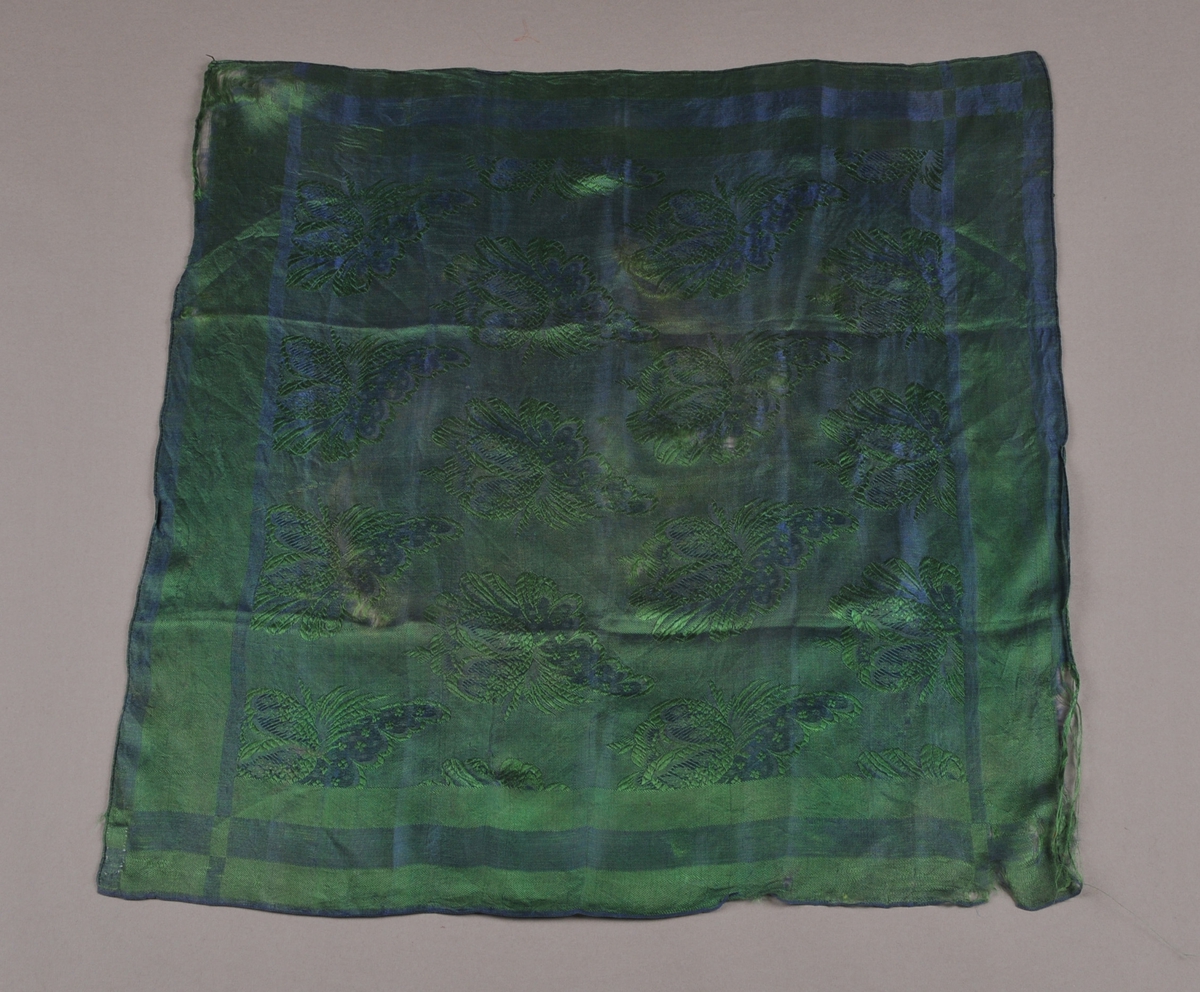 Damaskvove silketørkle i blått og grønt. Motiv: striper langs ytterkantane og roser og blad på midtfeltet. Smal (2mm) fald langs tre sider, jarekant langs ei side.