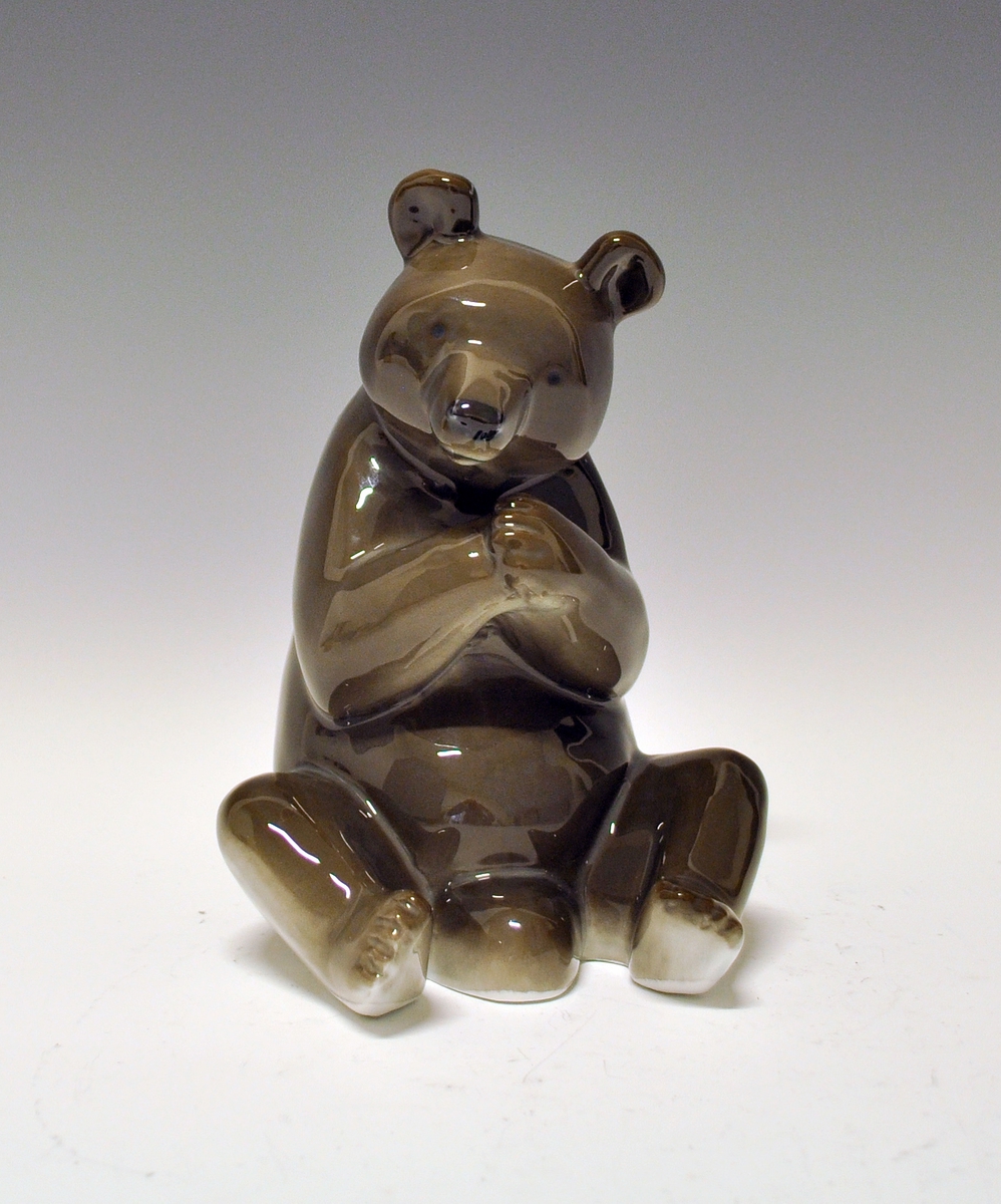 Figur av porselen. Sittende bjørn med forlabbene samlet under snuten. Brunfarget.