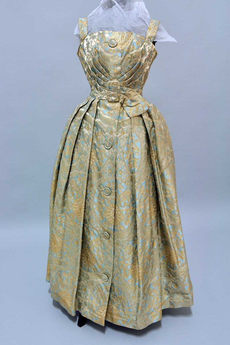 Selskaps- eller gallakjole med fast underskjørt i tyll. Stropper over skuldrene. Glidelås bak i ryggen. Knapper og beltespenne trukket i samme stoff som kjolen.