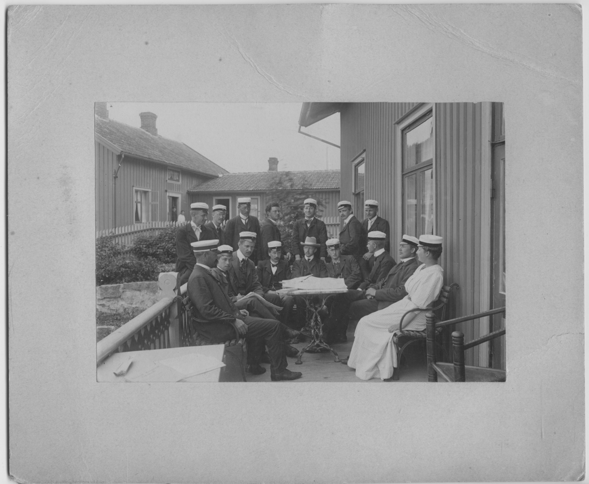 'Kristinebergs zoologiska station: :: Gruppbild, 9 personer, 8 män och 1 kvinna sittande vid 1 bord på veranda. 7 män stående bakom. Alla iklädda studentmössa utom 1 ung man utan huvudbonad och 1 man med hatt. I bakgrunden tittar 2 personer ut genom dörr.'