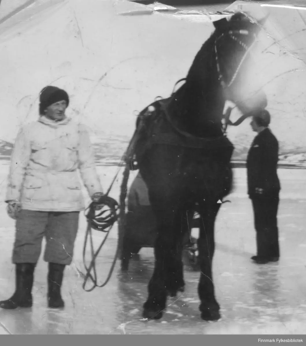 Johannes Nymo ute på havisen ved Brennelv i Porsanger med hesten Virda. En delvis skjult person sitter i sleden og en ukjent mann står til høyre på bildet.