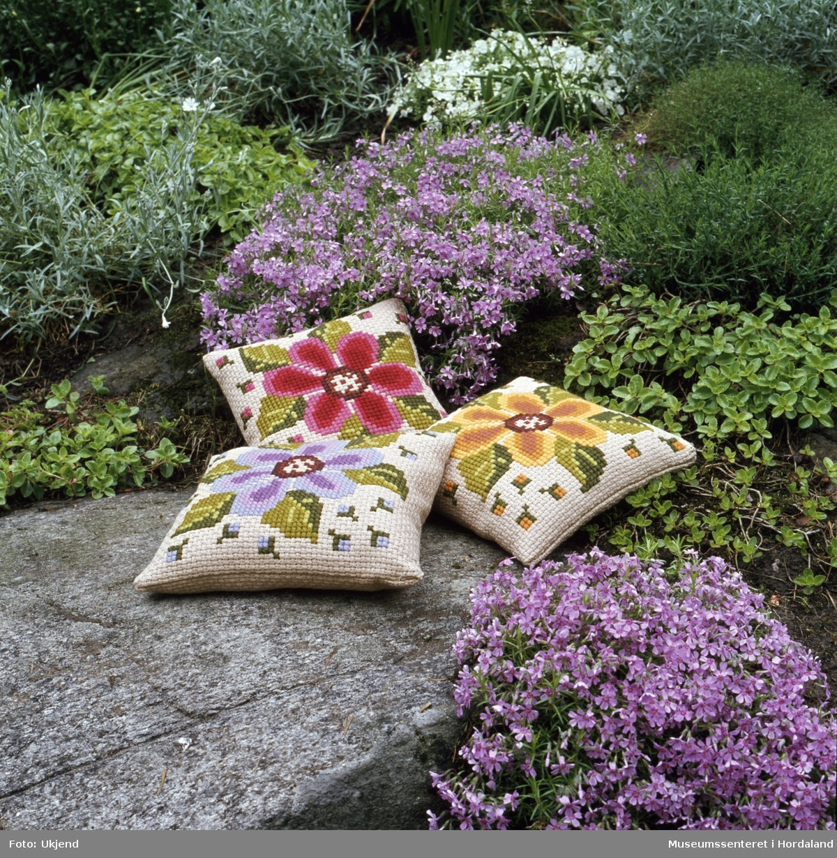 Reklamefoto av tre puter i blomsterbed av Design Gunnar P. Korssting i ullgarn på bomullsaida. Mønsterdesign: Laila Simonsen.
