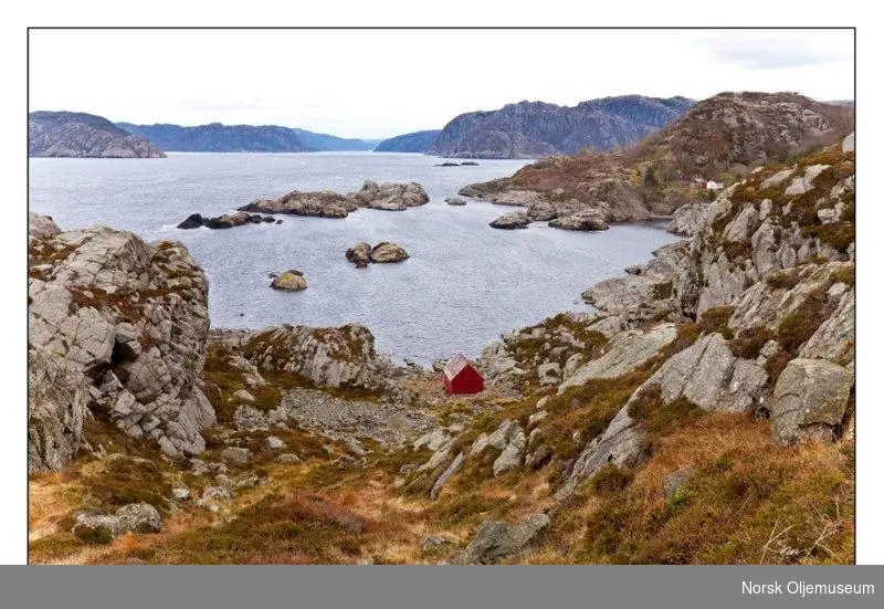 Fotografi av kysten på Lista med berg, holmer, øyer og hav. Midt i bildet ligger et lite rødt sjøhus.