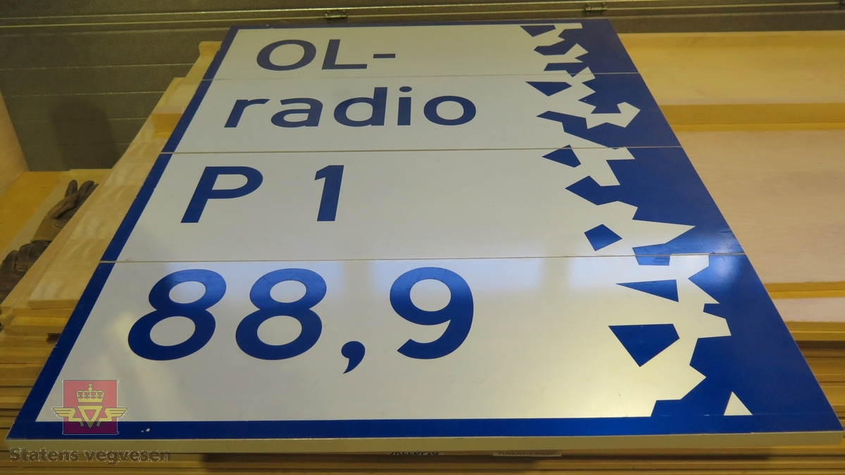 Rektangulært skilt av aluminium. Har teksten "OL-radio P 1   88,9". Hvit og blå, med grå bakside.