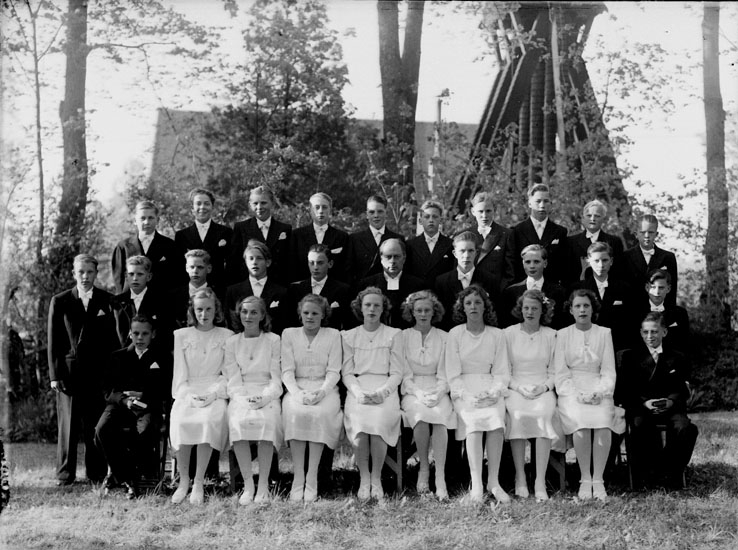 29 konfirmander, 8 flickor, 21 pojkar och prästen Bror Strömberg.
Almby kyrka och klockstapeln i bakgrunden.