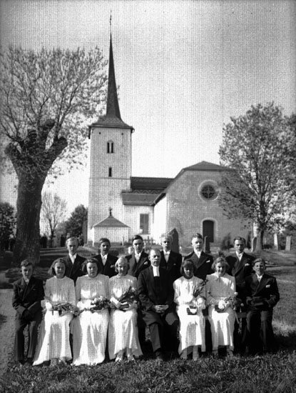 13 konfirmander, 5 flickor, 8 pojkar och kyrkoherde Gabriel Carlsten.
Gällersta kyrka i bakgrunden.