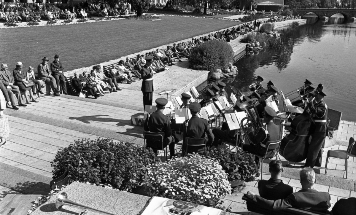 I 3 musikkår, 2 september 1965. 
Militär musikkår i Centralparken. Till vänster sitter publik utplacerade på ett flertal stora parkbänkar längs med vattnet. Människor sitter även på stenmuren samt på stolar på en utomhusservering längre bort. I bakgrunden skymtar en bro över vattnet.