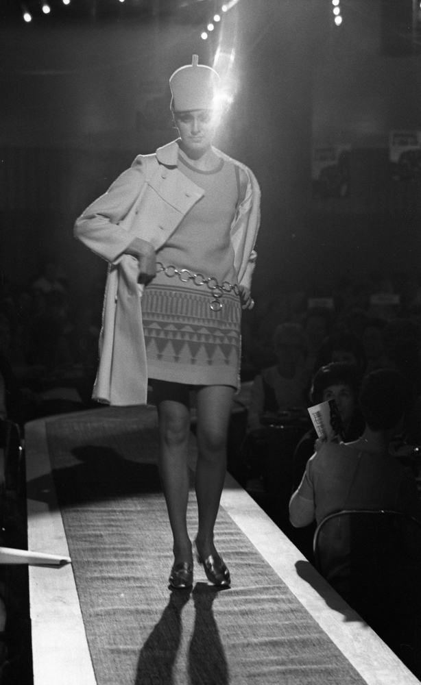 Modevisning, Görtz motor (Rep av oil) 6 april 1967

En fotomodell går på catwalken under en modevisning. Hon är klädd i en vit knappa. Den är uppknäppt och under den har hon en ärmlös lårkort klänning med ett metallskärp i midjan. Hon har en ljus hatt på huvudet samt silverskimrande skor. Publik sitter nedanför scenen och tittar på.