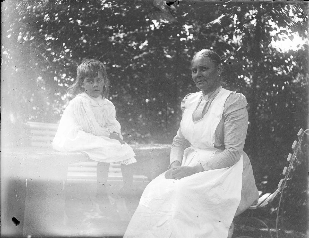 En kvinne sitter på en benk i en hage, på bordet ved siden av benken sitter en sommerkledt jente. Begge ser alvorlig på fotografen.