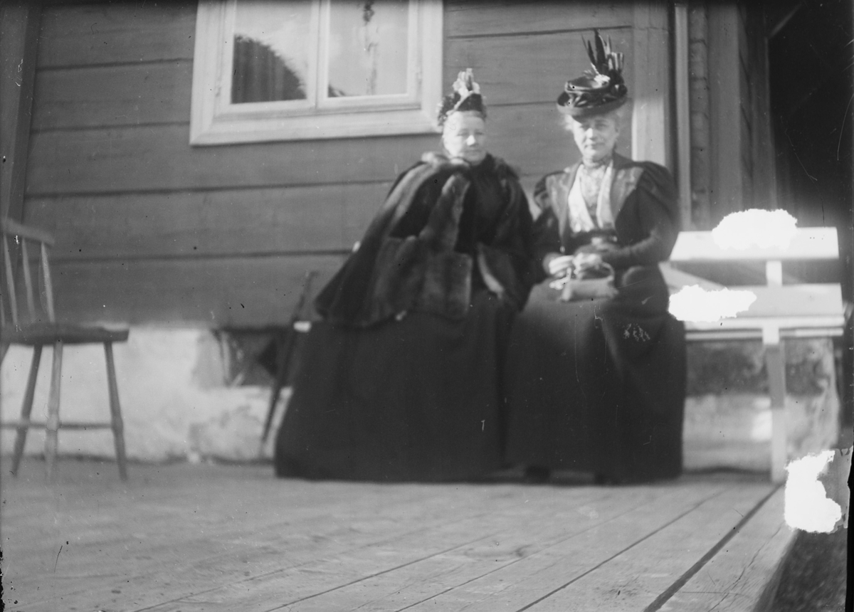 To damer, godt kledt, sitter sammen på en benk utenfor et hus. En vanlig kjøkkenstol står ved siden av dem på terrassen.