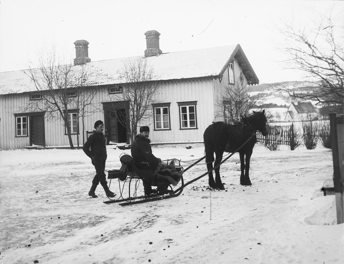I snøen står en hest med slede på tunet til Stiklestad Nordre. I sleden sitter en mann. En yngre mann kommer gående mot sleden. I bakgrunnen dominerer et gårdshus. Kirken i bakgrunnen er Stiklestad kirke.