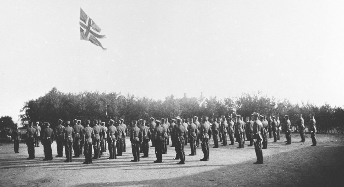 Vi antar at bildene er tatt i forbindelse med gjenopprettelse av Varangerbataljonen 01.07.1934. Bildet viser oppstilling på ekserserplassen på Nyborgmoen 1934. Soldatene står med ryggen mot kamera.