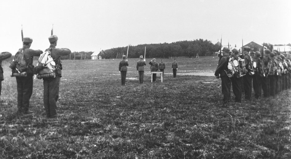 Vi antar at bildene er tatt i forbindelse med gjenopprettelse av Varanger batlajon 01.07.1934. Bildet viser soldater med full oppakning i givakt og offiserer på eksersersplassen på Nybergmoen 1934.