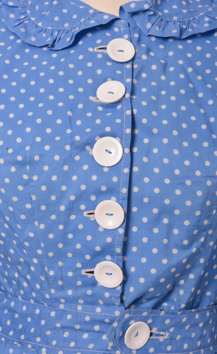 Sommerkjole m/hatt. a) Bomullstoff, blått med hvite prikker. Sammensydd i livet. Overdelen har Lukning foran med 5 hvite knapper og knapphull, puffermer. Krage med heppekant. 6 Bredder i utskrådd skjørt. Lukning til venstre for midt foran. 
b) Belte med knapp og knapphull. 
c) Hatt, Heklet av hvitt bomullsgarn. Ståltråd ytterst i Bremmen (rusten) Figurkant på pullen. Pyntebånd av kjoletøyet.