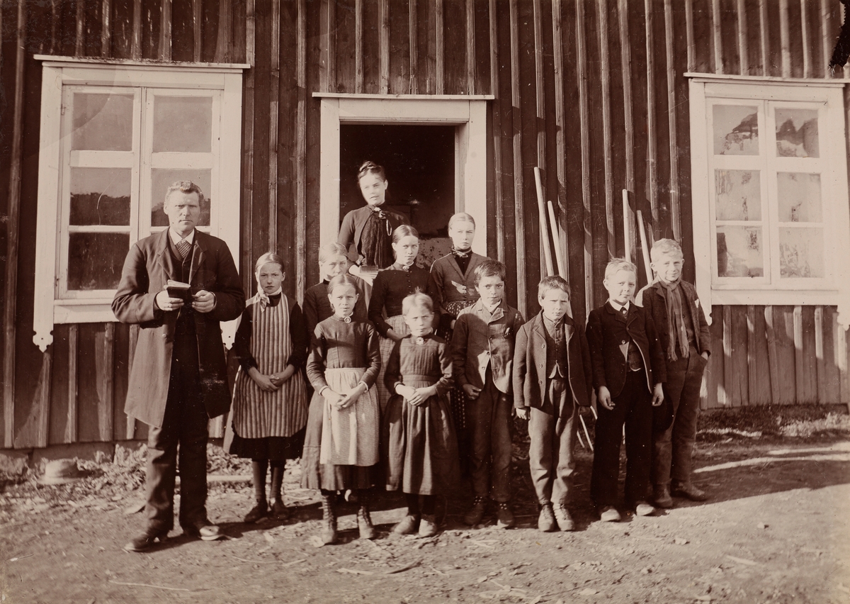 Gruppebilde fra Thorsø. En mann som holder en liten bok, syv jenter og fire gutter er stilt opp foran en dør.