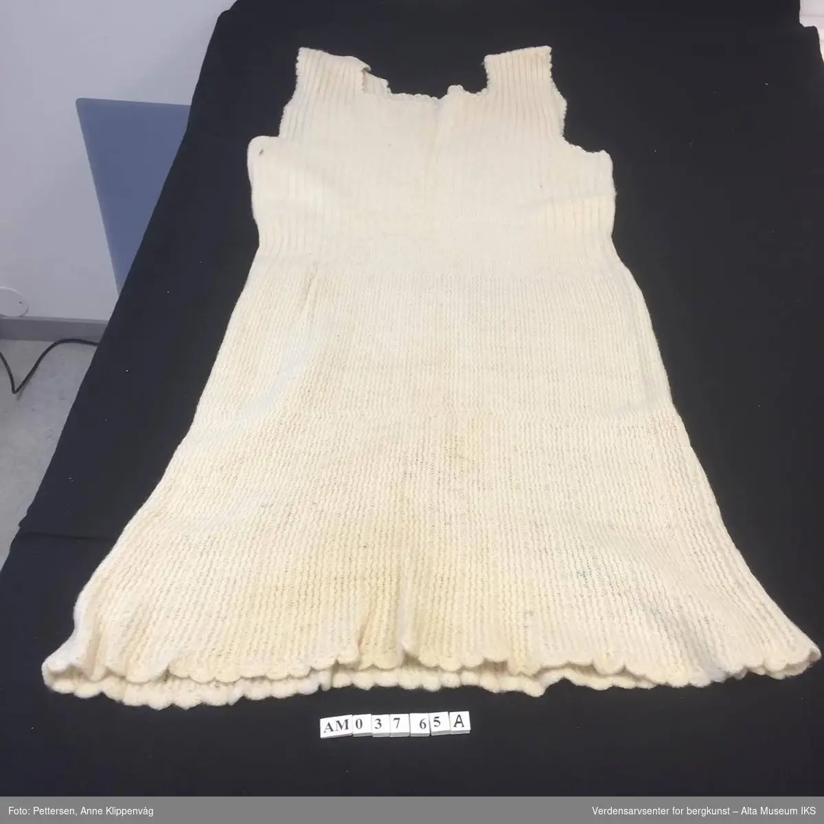 Tre produkter fra strikkemaskinen i samme gruppering:
AM.03765.A: Kjole. Hvitfarget og ermeløs kjole. Ribbestrikket mønster. Heklede kanter langs kjolens nederste kant. 
AM.03765.B: Et avklippet, hvitt gensererme. 
AM.03765.C: Et grått sokkpar.