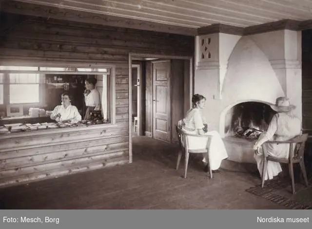 Interiör från Abisko turiststation. Ett hörn av Lillstugan, två kvinnor sitter vid öppen spis, två kvinnor bakom försäljningsdisken.