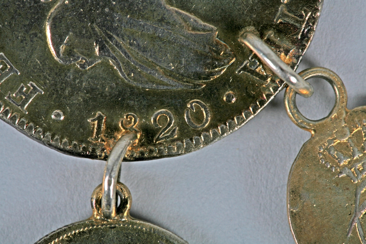 Sølvkjede satt sammen av tynne sølvringer. Anhenget består av en sølvmynt med diameter 39mm med tre mindre mynter festet på. Hver av de små myntene har et løv festet på. 2 av de små myntene er 15mm i diameter, den midterste er 17mm i diameter. 