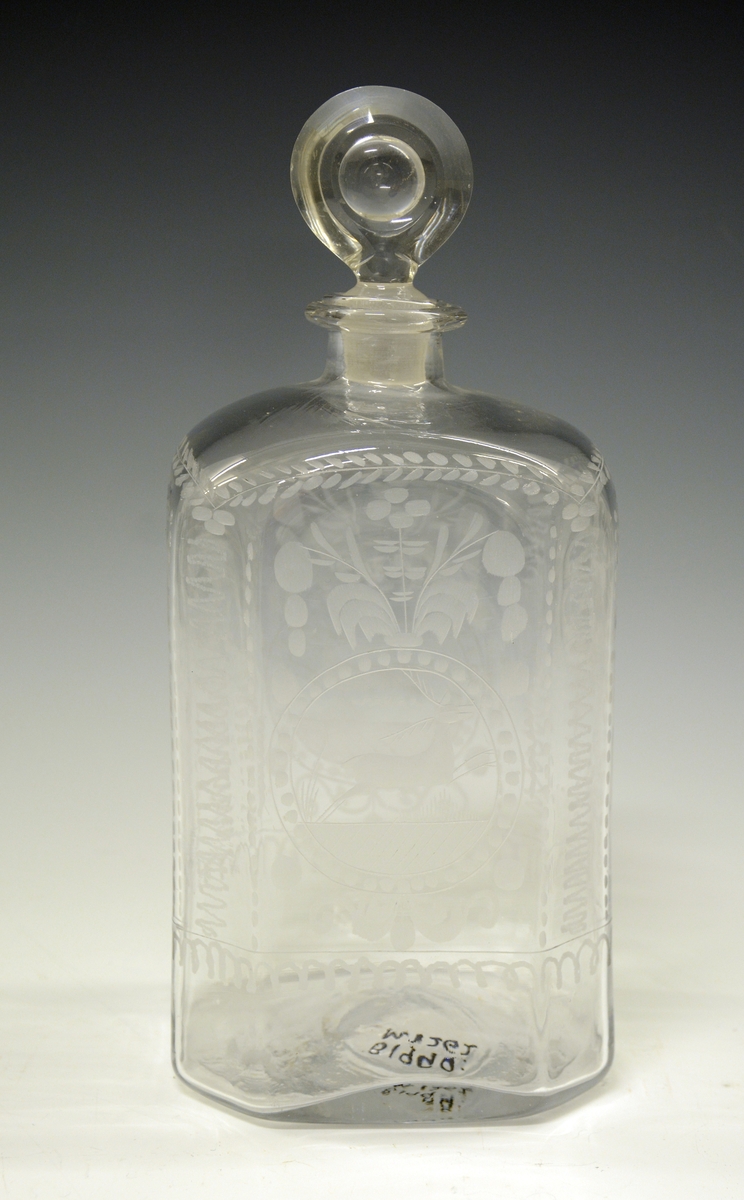 Glasskaraffel i klart glass - firkantet form, med avbrutte hjørner (skrå), slik at flasken eg. har 8 kanter. Gravering på alle sider - midt på begge de store flatsidene er det gravert et dyr (hjort eller rådyr), ellers vanlige ranker, blomster og blader.
Flasken har glasspropp - virker original. Ingen skader.

Stor glassflaske av typen kantineflaske. Gravert. Usikker produsent - kan det være Gjøvik eller Hurdal? 1790-1820 ca.