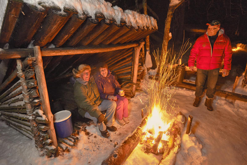 Bildet viser 3 personer samlet i og ved en gapahuk. Foran gapahuken brenner det livlig i et bål.