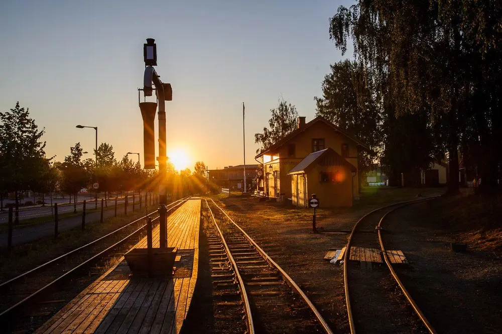 Urskog-Hølandsbanens stasjon på Sørumsand badet i sol. I forgrunnen vanntårnet, i bakgrunnen ligger stasjonsbygningen på høyre side, i midten perrongen og skinnegangen, og til venstre skimtes bilveien.