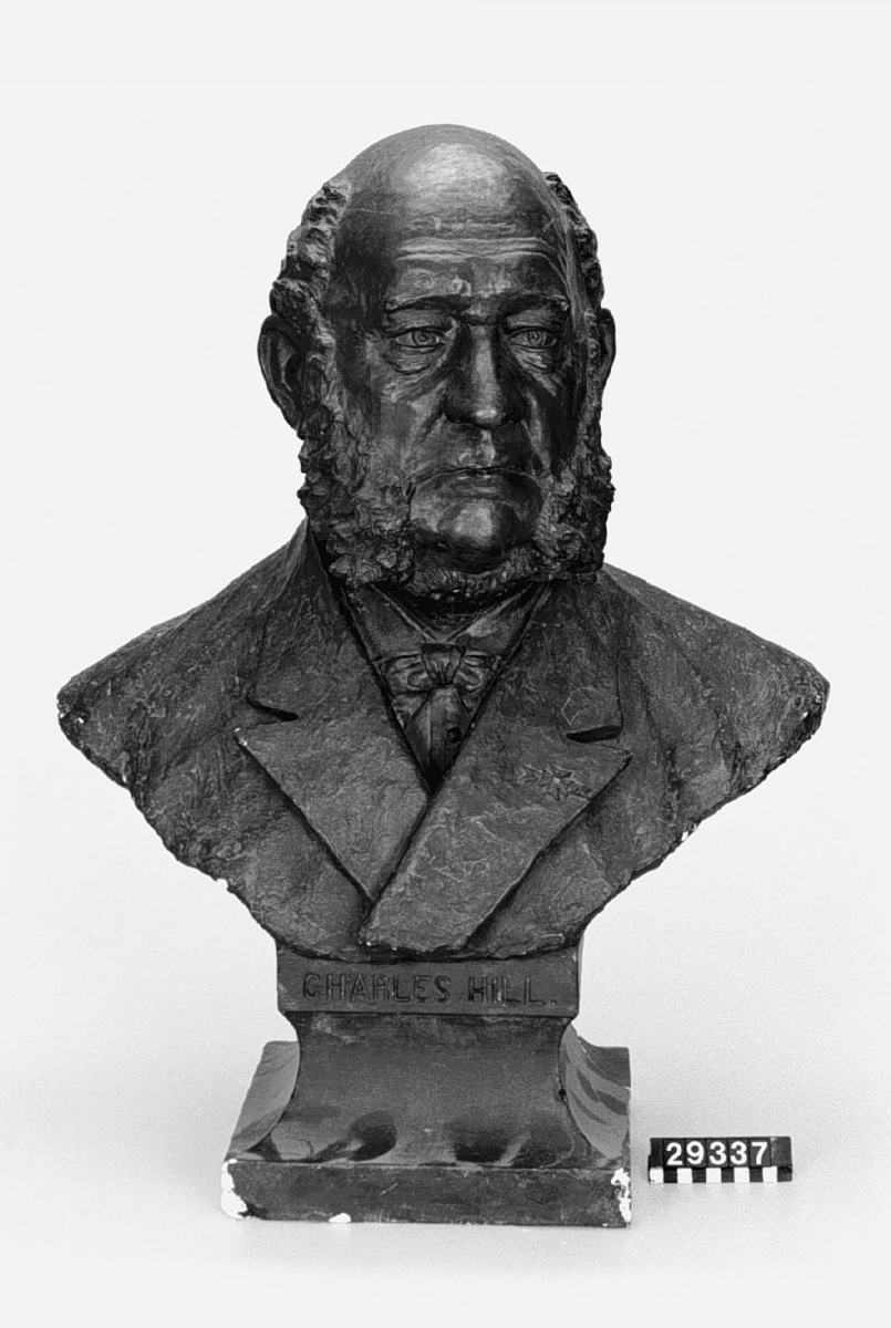Byst av "Charles Hill" (1817-1889). Utförd i bronspatinerad gips på marmorerad träpiedstal. Under bysten: Charles Hill. Sockelns baksida: Sc'Sven Andersson 1887.