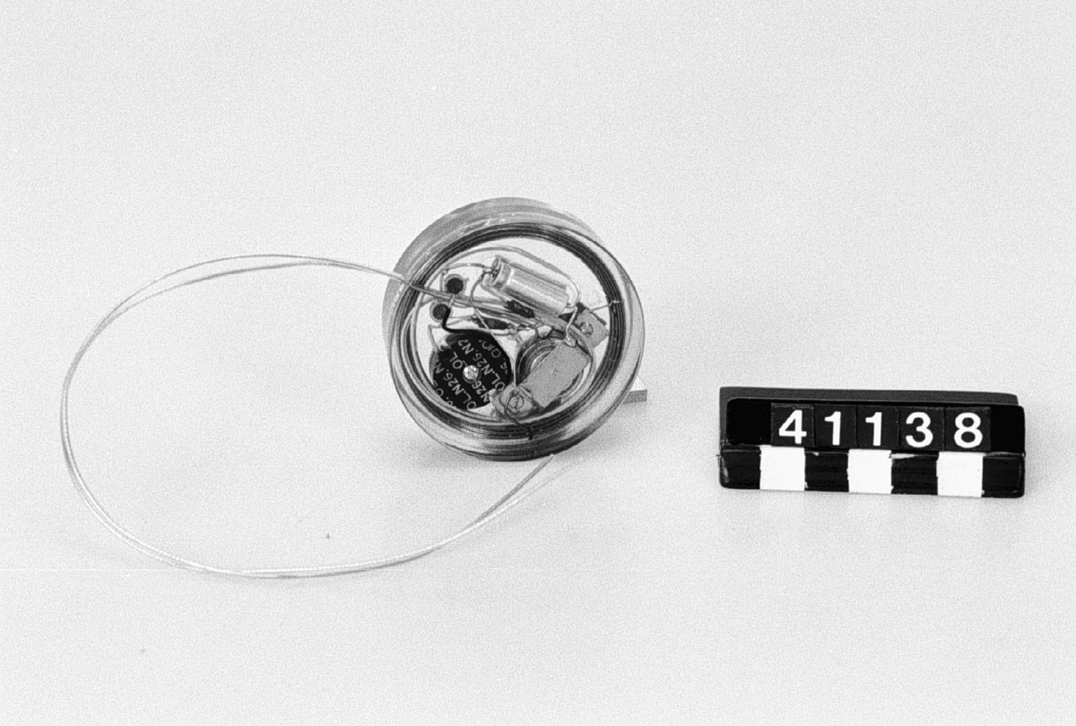 Pacemaker för implantat, med ett fåtal ingående komponenter. Ingjutet i kloss av epoximaterial med två elektroder för stimulans av hjärtverksamheten.
