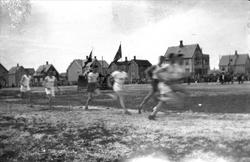 Løpere på et idrettstevne. Flagg, tilskuere og bygninger i b