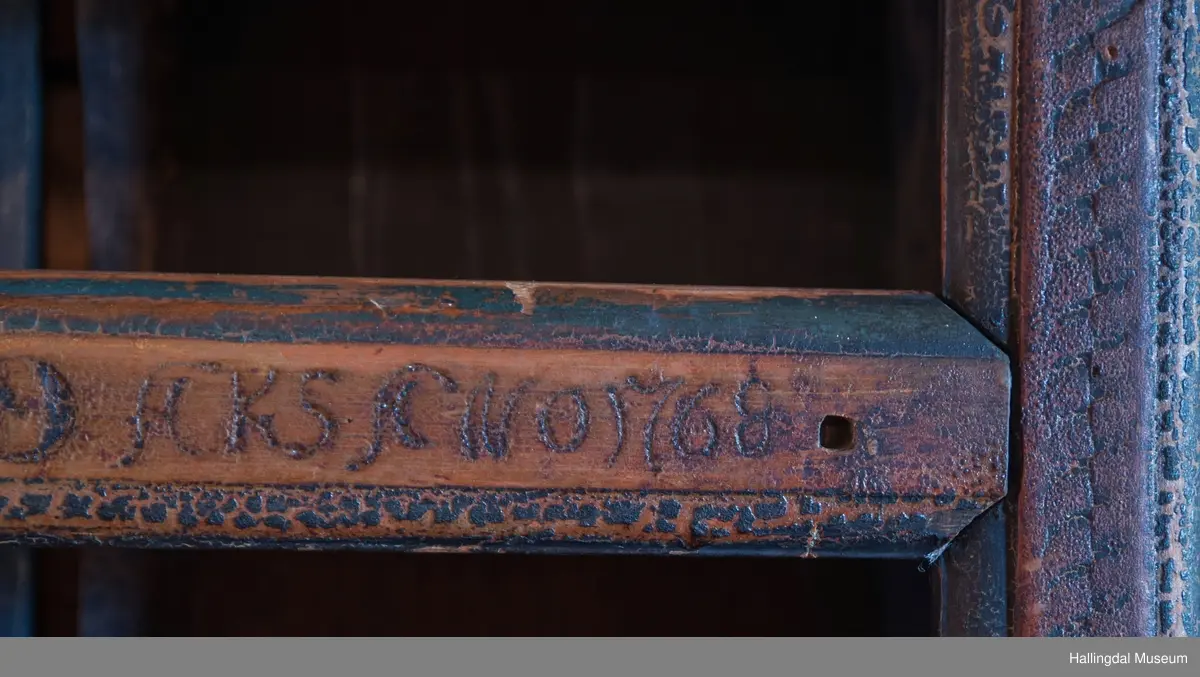 Rester etter rød og blå maling.  Gesims, skråner ned bakover. Profilert. 6 sirkulære hull i arkitraven.  Mellom disse er malt med sorte bokstaver.  Gavlene har utskårne "hylleknekter".  Bakveggen består av fire vertikale planker.  Skjøtene er dekket med profilerte lister.  Bare to biter i nederste del til venstre er fremdeles på plass.  Nederste del er lukket skap.  Tredelt, midterste del er skapdør.  Platen har profillist.  Feltene har speilfyllinger.  Blåmalte.  Klosset marmorering.  Speilfyllingenes form er som en liggen H.  Har profillist.  Skapet er satt på en ny grønmalt plate.  På døren er malt hvite bokstaver.  Nederst på døren er skåret inn bokstaver, opp-ned.  To nøkkelhull m. jernbeslag på venstre side av døren.  Over døren til venstre, en dreibar trekloss, holder igjen.  Innvendig har døren merke etter stort jernbeslag på samme side som nøkkelhullene.  To lister skåret i treverket minner om lange jernbeslag,-smalner mot nøkkelhullene.  Akantusranker i rødt, blått og hvitt, meget avslitt.  En hylle i skapet. Mangler bakvegg.  Karmen har på venstre side spor etter to jernhengsler.  Øverst på høyre kortside, mangler gesimsen en bit, og holdes til sideveggen med en jernkrampe.  Jernkrampe også på venstre side.  Skapplaten mangler list på høyre side.  Venstre side, fra gesimsen og ned har spor etter å ha stått inntil en vegg eller et annet møbel.