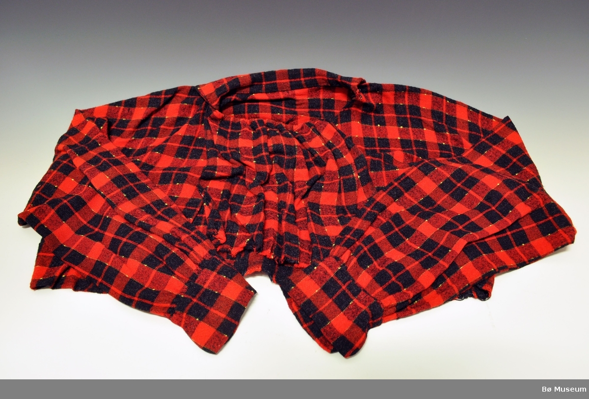 Skjorte med krave og brystduk, i rødt og svart rutemønster. Skjorten er sydd i et stykke tøy, det er klippt ut til ermer i hovedtøyet.