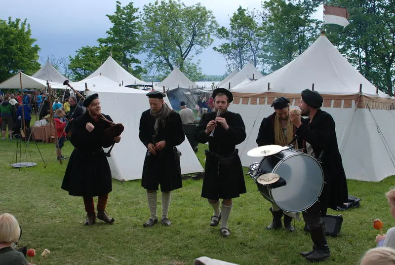 Four men in black medieval cotehardies playing bagpipem shawmsm and drums